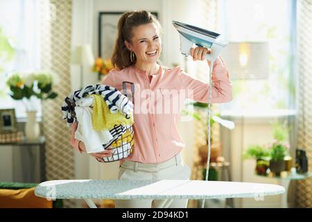 Portrait of smiling stylish female in pink shirt and white pants with iron and washed clothes basket in the modern house in sunny day. Stock Photo