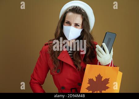 Hello autumn. Portrait of elegant housewife in red coat with smartphone and paper shopping bag on brown background. Stock Photo