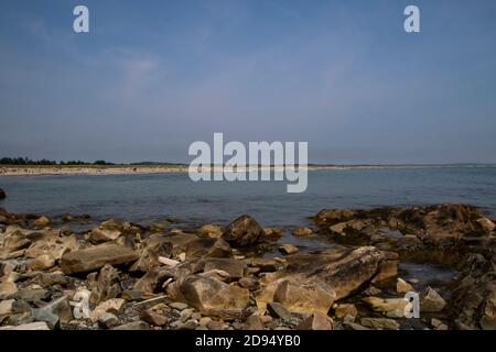 A view of a people filled beach as seen from across a bay with a rocky shoreline in the foreground.  The blue sky has an number of wispy clouds in it. Stock Photo