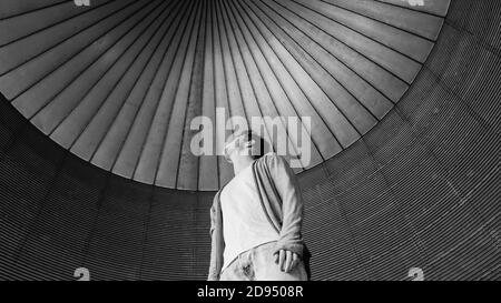 Grayscale shot of a young male wearing a white t-shirt paired with jeans and a jacket in a building Stock Photo