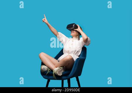 Lovely woman sitting in armchair is pointing at the free space while testing a vr headset on a blue wall Stock Photo