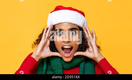 Shocked woman in Christmas attire yelling with hands cupped around face on yellow isolated studio background Stock Photo