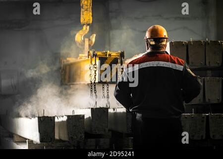A male worker controls the production process in a factory as a crane moves a reinforced concrete product with holes. Reinforced concrete pillars secu Stock Photo