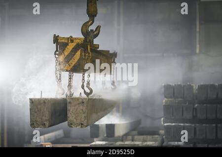The crane moves a reinforced concrete product with holes. Reinforced concrete pillars fixed with metal hooks and chains on the background of the plant Stock Photo