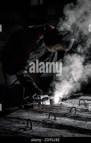 A young  man welder in  uniform, welding mask and welders leathers, weld  metal  with a  welding machine at the construction site, white smoke Stock Photo
