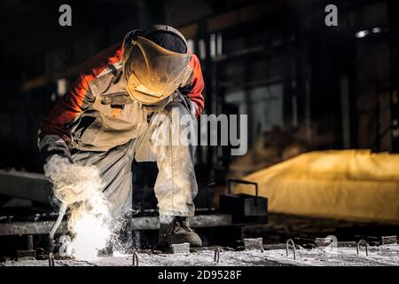 A young  man welder in  uniform, welding mask and welders leathers, weld  metal  with a  welding machine at the construction site, yellow sparks fly t Stock Photo