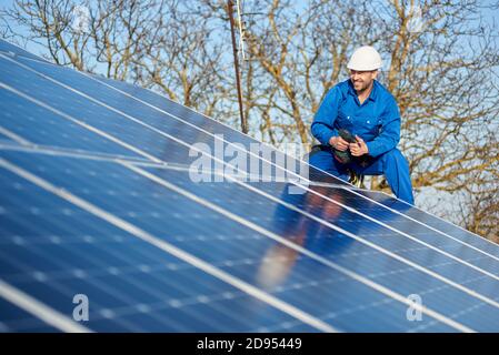 Smiling male engineer in blue suit and protective helmet installing photovoltaic panel system. Electrician mounting solar module on roof of modern house. Alternative energy concept. Copy space Stock Photo