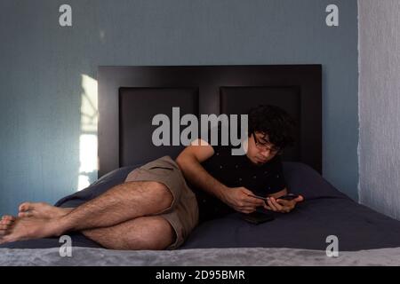 Hispanic young man lying on a bed shopping online with his smartphone and a credit card on his hand Stock Photo