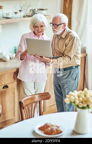 Loving older family spouses standing at home and looking at computer screen while showing video. Lifestyle concept Stock Photo