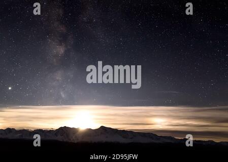 Amazing night landscape with starry sky, Milky Way galaxy, moon and beautiful clouds on the horizon above snowy mountains Stock Photo