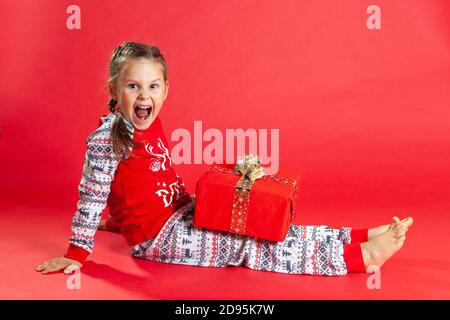 laughing girl with two pigtails and in Christmas pajamas sits on the floor and holds a gift box on her lap Stock Photo