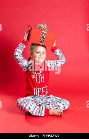 smiling girl with two pigtails and in Christmas pajamas holds gift box above her head, isolated on a vibrance background Stock Photo