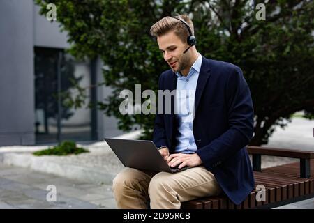 Portrait of handsome smiling man in casual wear sitting on bench using laptop for online meeting, video call, video conference. Communication online Stock Photo