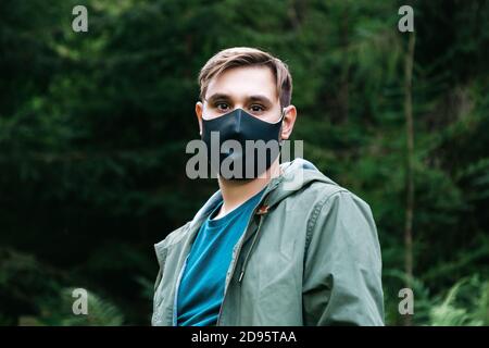 Caucasian man in black protective mask outside. Guy portrait in medical mask on forest landscape alone keeping social distancing. Protection against Stock Photo