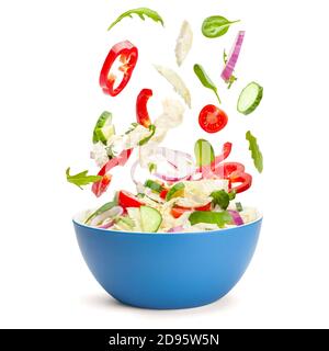 Vegetable salad in levitation. Fresh vegetarian salad of cucumbers, cabbage, cherry tomatoes, onions, peppers, spinach and arugula. Flying pieces of salad over a blue saucer isolated on white. Stock Photo