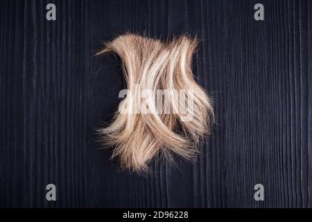 Blond wavy hair lock black wooden background closeup, cut off natural blonde hair curl on dark wood, haircut, hairstyle, human hair clipping hair snip Stock Photo