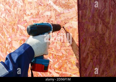 A gloved man's hand holds a screwdriver and fixes a wooden panel to the wall. Stock Photo