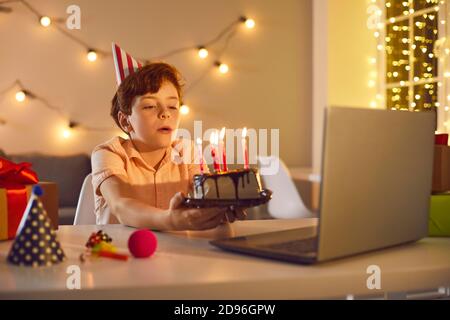 Little birthday boy making a wish and blowing burning candles on cake during online party at home Stock Photo