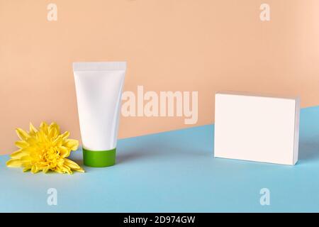 White cosmetic tube with green cap and no label next to yellow chrysanthemum flower and box against colorful studio background. Close up, copy space Stock Photo