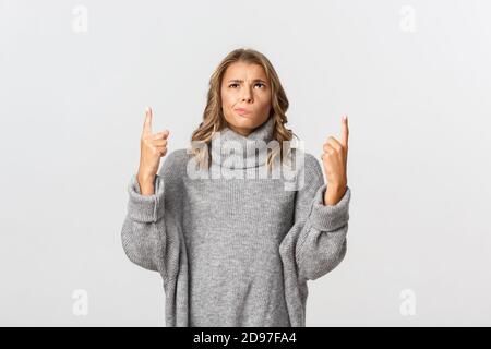 Image of skeptical blond girl in grey sweater, looking indecisive and pointing up, smirking doubtful, standing over white background Stock Photo