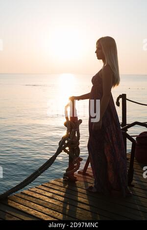 Blonde woman in a long sundress walks by the sea at dawn Stock Photo