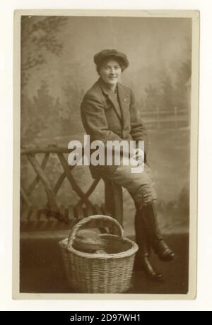 Original WW1 era studio postcard of attractive Land Girl in the Women's Land Army (WLA) - wearing breeches, jacket and hat with a basket of bread loaves, involved in harvesting wheat, agricultural section of WLA, dated August 1918 on the reverse, U.K. Stock Photo