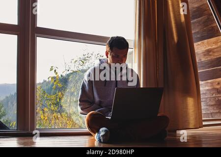 Man using laptop sitting near big window in wooden cottage Stock Photo