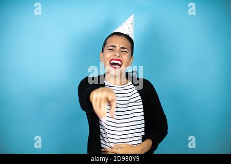 Young beautiful woman wearing a birthday hat over insolated blue background laughing at you, pointing finger to the camera with hand over body, shame Stock Photo