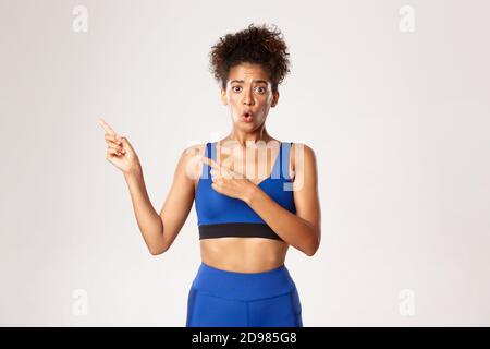 Studio shot of worried african-american female athlete in blue sportswear, looking concerned and pointing fingers left, standing against white Stock Photo