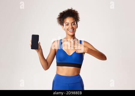 Sport and technology concept. Beautiful athletic woman in fitness clothing, pointing finger at smartphone screen, standing against white background Stock Photo