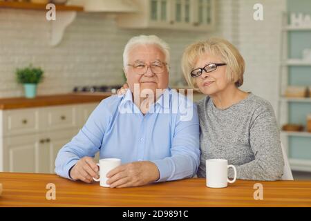 Portrait of happy senior couple sitting in the kitchen hugging and drinking tea or coffee. Stock Photo
