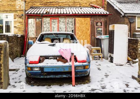 A bunch of remembrance flowers on the bonnet of a snow covered old broken down car at the rear of a boarded up house in Consett, County Durham, UK Stock Photo