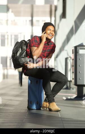 Portrait of happy travel man sitting on suitcase at station while talking on cellphone Stock Photo