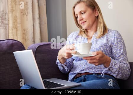 Mature Woman Sitting On Sofa Look At Laptop And Drinking Hot Drink Stock Photo