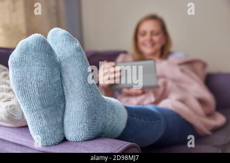 Woman At Home Wearing Cosy Warm Socks And Wrapped In Blanket Lying On Sofa Watching Digital Tablet Stock Photo