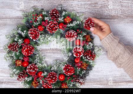 female hand in a knitted beige sweater adds a red painted bump to a homemade Christmas wreath on a wooden background Stock Photo