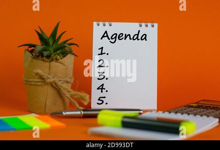 White note with inscription 'agenda' on beautiful orange background, colored paper, metalic pen, yellow marker, flowerpot with a plant and calculator. Stock Photo