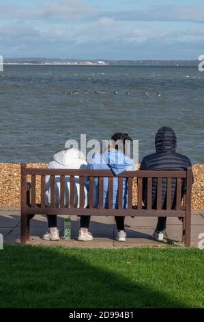 three youths sitting on a bench using mobile device on the seafront at the seaside together. Stock Photo