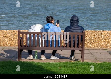 three youths sitting on a wooden bench at the seaside playing with mobile devices and phones taking selfies next to the sea. Stock Photo
