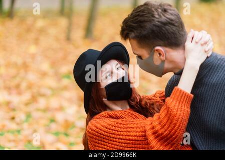 Young red haired woman puts on a face mask while walking with young man in autumn park. Stock Photo