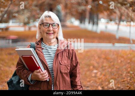 Smiling adult woman in casual clothes stands holding notebooks and backpack walking in the autumn park Stock Photo