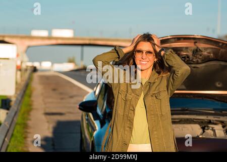 Stressed young woman standing near her broken car on the highway roadside. Stock Photo