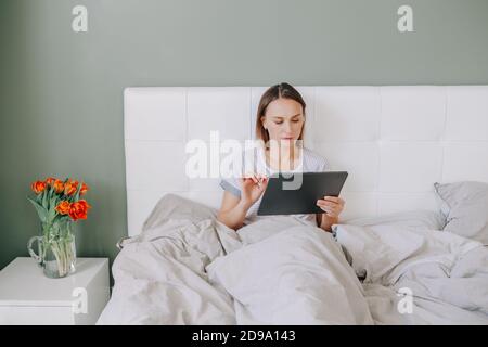 Beautiful middle age woman sitting in bed in morning with digital tablet surfing Internet. Young woman talking with friends in social media. Stock Photo