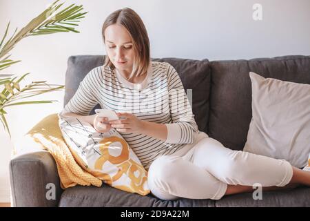 Young woman sitting on couch and texting talking at social media on smartphone. Middle age woman on sofa typing messaging on mobile phone. Stock Photo