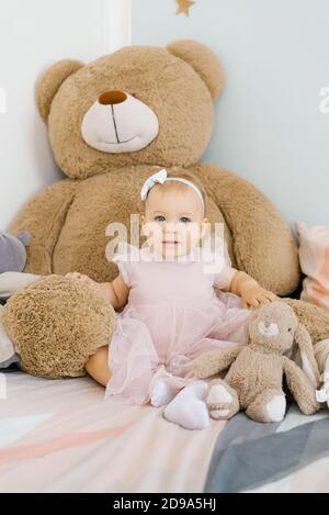 A cute one year old girl is sitting on the bed in the children's room at home among soft funny toys in a pink dress Stock Photo