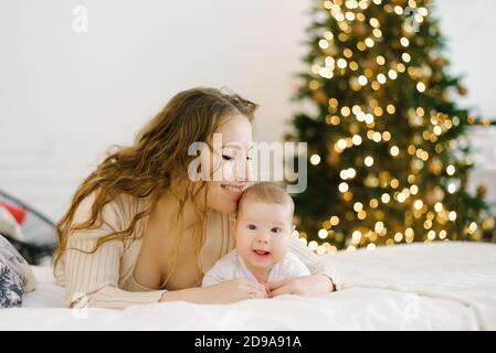 Happy mother lies on the bed next to her baby in the background of Christmas lights Stock Photo