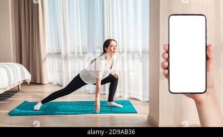 Yoga in the bedroom. A young pretty woman trains at home on a sports mat. A females hand holds a smartphone. Mock up. Side view. The concept of home w Stock Photo