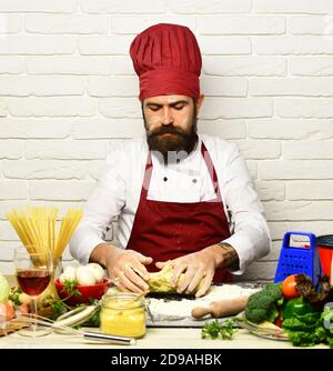 Restaurant cuisine concept. Chef makes dough. Cook with serious face in burgundy uniform sits by table with vegetables and kitchenware. Man with beard kneads dough on white brick wall background. Stock Photo