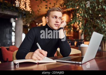 Successful young guy sits in cafe by table with his laptop on it Stock Photo