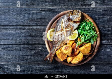 Whole roasted dorado fish with skin on baked potato coated in breadcrumbs served on a wooden cutting board with lemon wedge and fresh arugula or wild Stock Photo